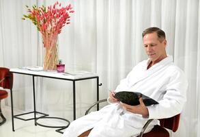 Handsome mature Caucasian man dressed in white terry bathrobe relaxing in private room of a wellness spa centre, sitting on an armchair and reading magazine photo