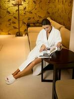 Beautiful relaxed middle aged woman in white terry bathrobe sitting on armchair in the lounge zone of a luxury wellness spa center and reading magazine between receiving body care beauty treatment photo