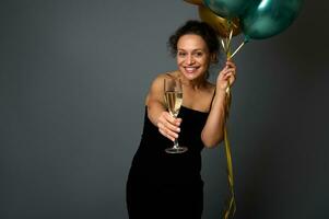 Cheerful woman celebrates event holds luxury shiny golden green air balloons and shows a flute with champagne to the camera holding it in outstretched hand. Birthday, Christmas, New Year concept photo