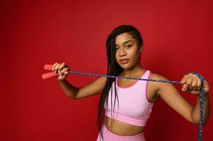 africano americano bonito mujer en rosado ropa de deporte participación un saltando cuerda en frente de su y demostración eso a el cámara con confianza mirando a él, posando terminado rojo antecedentes foto