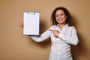 Smiling woman pointing her index finger on white blank paper sheet on clipboard, standing against beige background with copy space photo