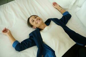 High angle view of a delighted pretty businesswoman in business casual suit lying down on a bed in bedchamber of a hotel room and smiles toothy smile looking up photo