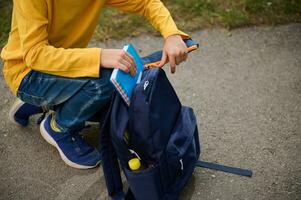 Close-up, cropped view of a schoolboy putting a notebook and pencil case in a backpack, returning home after shooting. Back to school concept photo