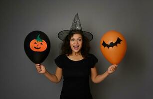 Surprised woman wearing a wizard hat poses with black and orange air balloons with felt-cut bat and pumpkin, looking at camera. Halloween concept on grey background, copy space for advertising photo