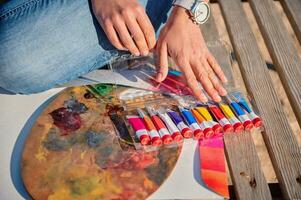 Close-up of hands on a painting palette and tubes with paints on a wooden chaise lounge photo