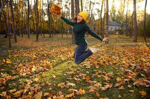 Beautiful woman in warm green pull-over, yellow woolen and casual denim hat jumping high with a bouquet of beautiful dry autumn maple leaves in hands, enjoying beautiful autumn day at sunset in park photo