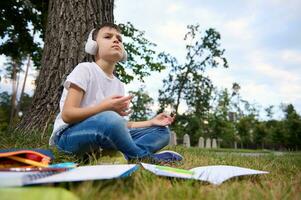 Adorable school child boy feels exhausted and tired after school and homework, sits in lotus position and meditates with wireless headphones on head. Workbooks and school supplies lying on the grass photo