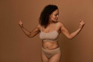 Delighted confident middle aged African American woman with stretch marks, in beige underwear looking at her arms and clenched fists, posing against colored background with copy space. photo