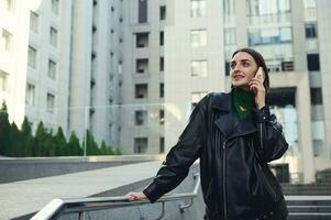 Attractive brunette woman with stylish short hair talking on mobile phone standing outdoor against modern corporate high-rise buildings. Digital gadgets and communication concepts in contemporary life photo