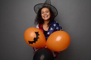 Attractive happy mixed race woman in wizard hat, wrapped in American flag, holds colorful orange air balls, smiles toothy smile looking at camera. Halloween concept on gray background with copy space photo