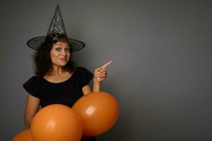 Cheerful joyful happy Hispanic woman in wizard hat, dressed in black, holds orange inflated air balloons and points on a copy space on gray background. Halloween party concept photo