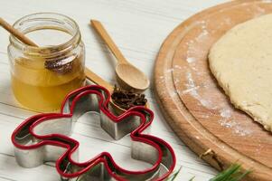 Cropped image of rolled dough on wooden board, man shaped gingerbread cutters and glass jar of honey on white wooden table photo