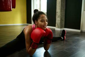 africano americano hermosa atlético mujer, hembra Boxer en rojo boxeo guantes estiramientos el piernas en el enroscarse en el piso de un Deportes gimnasio foto