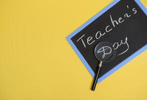 Flat lay composition from a magnifier, lens, loupe on a chalkboard with lettering Teachers Day on yellow background with copy space for text. photo