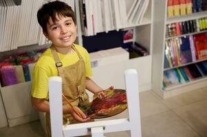 hermoso primario colegio chico, pequeño artista sostiene un paleta con petróleo pinturas, pintura en lienzo, sonrisas mirando a cámara foto