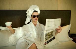 Portrait of a stylish woman in sunglasses and white terry bathrobe and head wrapped in towel, looking at camera resting after shower on the bed, holding cup of hot drink and newspaper in her hands photo