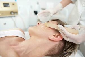 médico cosmetóloga aplicando gel con anestésico a mujer cara para preparando eso a láser pelo eliminación procedimiento en equipado spa clínica foto