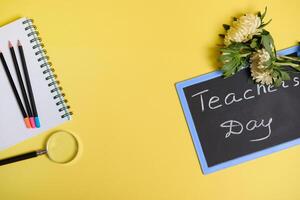 Flat lay of asters flowers lying down on chalkboard with text Teachers Day , and a magnifier loupe next to colored pencils on a blank sheets of a notepad isolated on yellow background with copy space photo