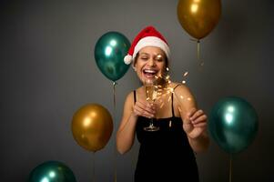 Charming gorgeous woman in Santa hat celebrates Christmas, rejoices lighting sparklers, standing with a glass of sparkling wine on a gray background decorated with beautiful gold and green balloons photo