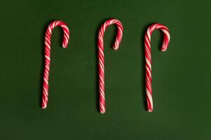 Flat lay Christmas composition of three stiped white and red sugary sweet lollipops candy canes on dark green background with copy space. Food composition with two contrasts colors photo