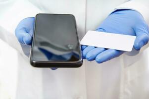 Cell phone and plastic white card on hands in medical gloves on the background of doctor's white uniform. Copy space. Health insurance concept photo