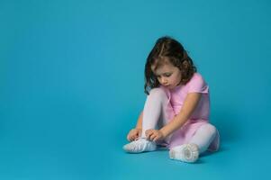 Cute ballerina focused on tying shoelaces on ballet shoes sitting on blue background photo