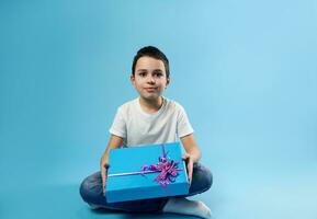 Front view of a cute smiling boy posing with a birthday gift in his hands on blue background with copy space. photo