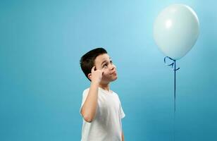 Little boy putting his finger on his temple and looking at the flying blue balloon with a thoughtful look photo