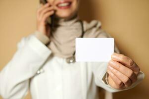 Closeup of a plastic blank white card on the hand of a female Muslim doctor wearing a hijab talking on the phone. Copy space for promotions. Advertising. Insurance concept photo