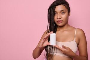 hermosa africano mujer con elegante afro rastas en beige ropa interior sostiene un tarro de hidratante y y demostración eso a el cámara. cuerpo y piel cuidado concepto. rosado antecedentes con Copiar anuncio espacio foto