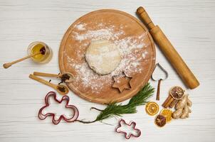 Dough on a round wooden board next to a rolling pin and kitchen utensils for making Christmas gingerbread cookies, cutting molds and dried orange slices and pine branches on a wooden surface photo