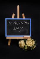 Close-up of a chalk blackboard with lettering Teacher's Day, standing on a wooden table easel, next to black alarm clock, isolated on black background with copy space. photo