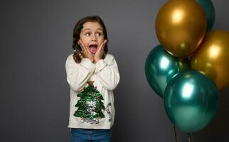 contento sorprendido bebé niña en suéter con brillante Navidad árbol, alegría en pie cerca inflado dorado y verde metálico aire globos en contra gris antecedentes con Copiar espacio. nuevo año concepto foto