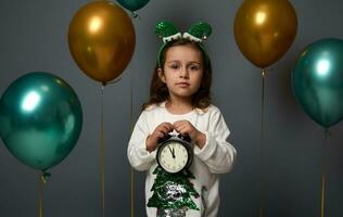 Adorable little girl wearing an elf hoop holds an alarm clock with midnight on the dial, looks at camera posing over gray background decorated with golden green air balloons. Merry Christmas concept photo