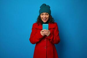 Isolated portrait on blue background of a cheerful awesome happy pretty woman wearing bright red coat and warm green woolen hat holding mobile phone and rejoices looking at camera. Copy ad space photo