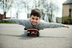 A cute boy lying with his belly on a skateboard with his arms outstretched like wings and imagining a flight photo