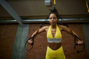 Determined slim body African woman with flat belly doing push-ups while exercising with suspension straps at gym. Healthy and active lifestyle, body care, fitness and sport concept photo