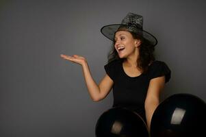 Joyful mixed race pretty woman dressed in witch carnival costume and wizard hat holds black air balloons, points on a copy space on gray background. Concept of Halloween traditional seasonal event photo