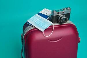 Studio shot of a suitcase, vintage camera, passport with air ticket and boarding pass, medical mask isolated over blue background with copy ad space. Tourism and travel concept photo