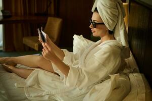 lado retrato de un hermosa mujer, negocio persona, envuelto en bañera toalla después relajante baños y disfrutando leyendo periódico, sentado en un cama en el hotel dormitorio durante su negocio viaje foto