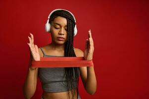 Determined sporty African woman with wireless headphones in gray sportswear stretching rubber band with arms while training, isolated over red background with copy space photo