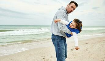 Handsome father holding his little cute daughter in his arms and playing together on the beach on a beautiful cool early summer day. Father's Day concept photo