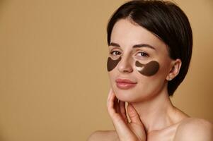 Headshot of an attractive Caucasian woman with beautiful healthy glowing skin looking at camera, posing against beige background with smoothing collagen eye patches under eyes. Copy ad space photo