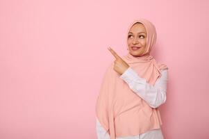 Gorgeous Arab Muslim woman with covered head in hijab looking at the pink background and pointing her index finger indicating the copy space for text. Business concepts with religious beautiful lady photo