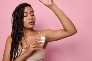 belleza retrato. de cerca de africano mujer con rastas en beige ropa interior aplicando antitranspirante debajo el axilas pureza, higiene, cuerpo cuidado concepto. foto