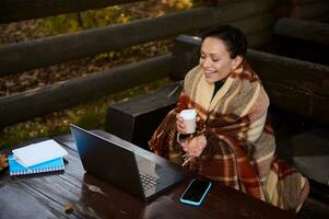 African American woman warming herself wrapped in plaid blanket and drinking hot coffee from paper cup during video call while sitting on open country terrace on cool autumn day in an oak grove photo