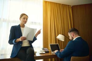 Self-confident European female boss holding a contract and reading text to her business partner, working on laptop in hotel room. Business concept with copy space for advertisement photo