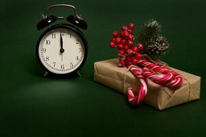 Close-up of black alarm clock with midnight on the dial, sweet striped lollipops, candy canes and holly on Christmas gift in craft wrapping paper isolated on green background with copy space for ad photo