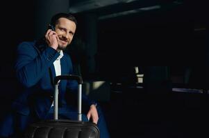 Handsome successful businessman in business suit smiles talking on cellphone, sitting in an armchair next to his suitcase in a lounge cafe of a luxury hotel. Young business investor at business trip photo