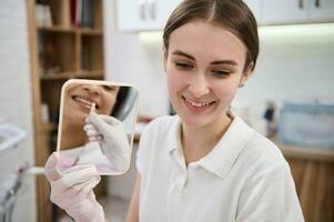 Headshot of beautiful female doctor, dental hygienist holding mirror with reflection of patient comparing shade of her teeth with dental sample color chart at dental clinic. Teeth whitening concept photo